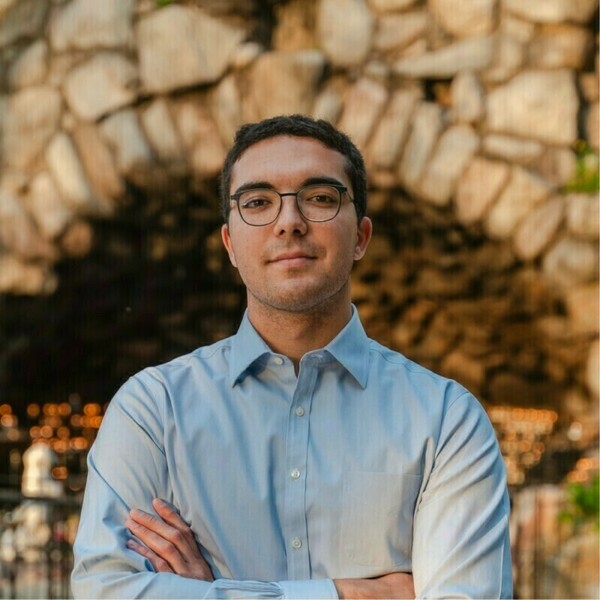 Young man looking at the camera with his arms crossed. He is wearing dark rimmed glasses, a light blue button down shirt, and has dark curly hair. He is standing in front of a stone structure.