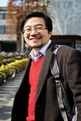 A smiling man with glasses, wearing a dark jacket over a red v-neck sweater, stands near a row of yellow bicycles.  He wears a gray backpack over his right shoulder. Photo of Silvano Mo Cheng, Peiking University.