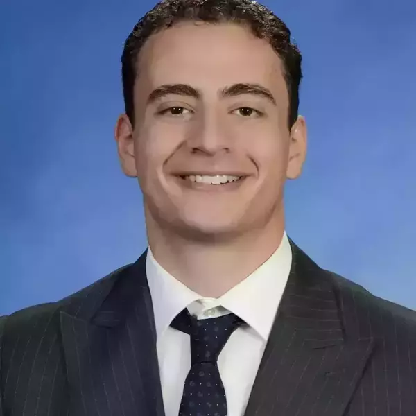A young man with short curly dark brown hair in a dark suit with a dark colored tie standing in front of a blue background smiling at the camera.