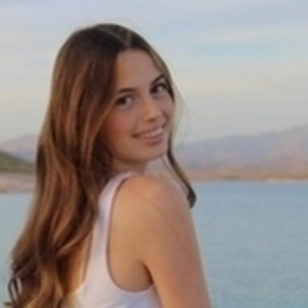 Young woman with long brown hair in a white tank top smiles over her shoulder, with a lake and hills behind her.