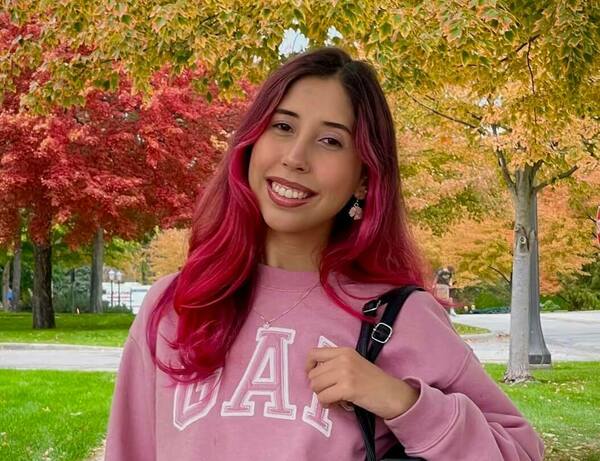 Young woman with vibrant pink hair smiles, wearing a pink GAP sweatshirt, against a background of red and gold fall trees.