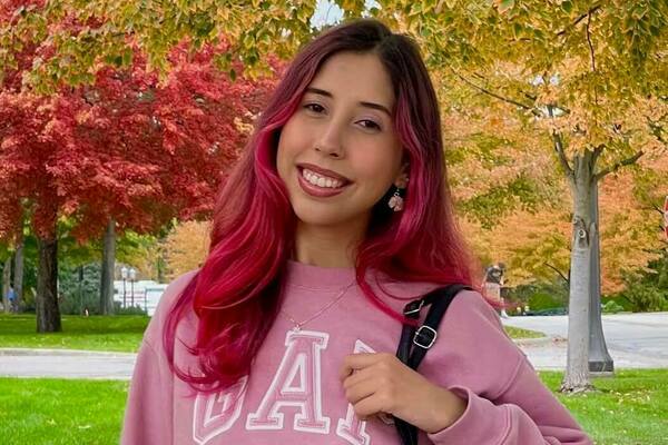 Young woman with vibrant pink hair smiles, wearing a pink GAP sweatshirt, against a background of red and gold fall trees.