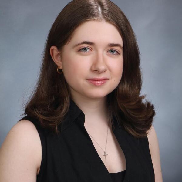 Young woman with long brown hair, blue-green eyes, gentle smile, in a black sleeveless top, gold hoop earrings, and cross necklace.