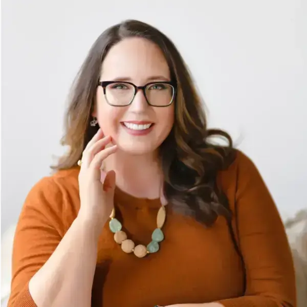Woman with long, dark, wavy hair, glasses, and a rust-orange top smiles, touching her cheek. She wears a green and tan beaded necklace and is set against a white background.