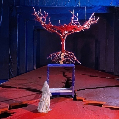 A red, bare tree sculpture with visible roots stands on a blue bed frame on a cracked red stage with a white-robed figure.